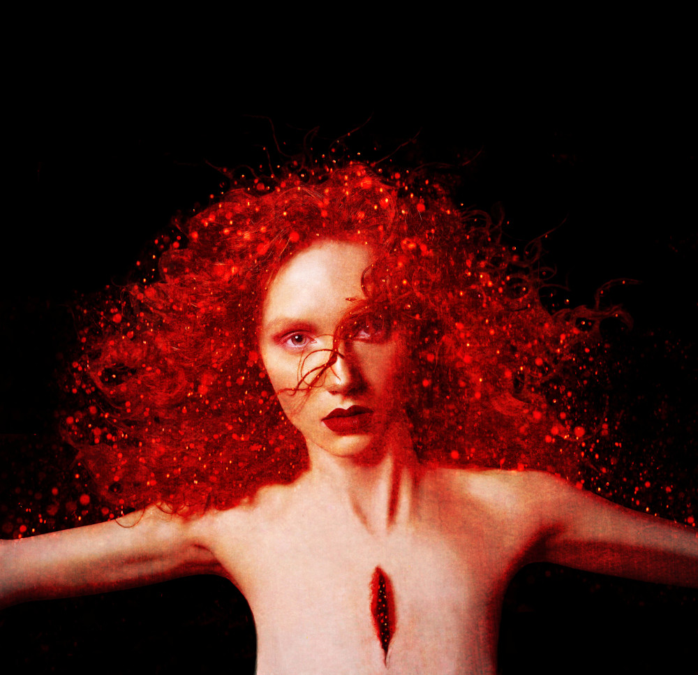 A woman with vibrant red, curly hair stares intensely. Her skin is pale, with a dark, deep gash on the chest.